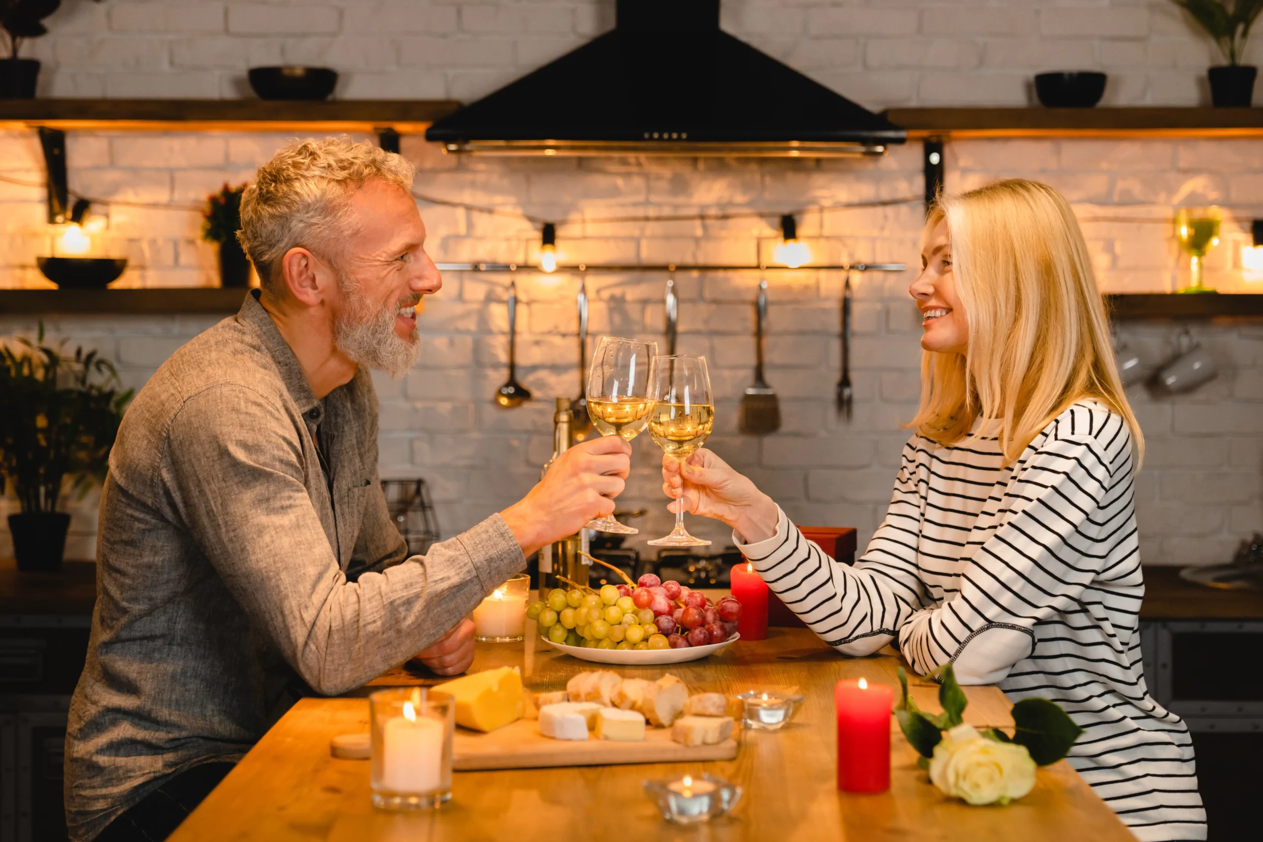 pareja brindando con una copa de vino blanco en una cocina sobre una mesa con diferentes alimentos